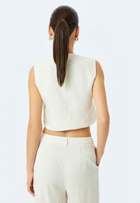 Beige, ärmelloses, verkürztes Oberteil mit glatter Textur, dezentem Stich und offener Rückseite, kombiniert mit dazu passenden hoch-taillierten Hosen.