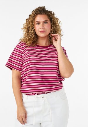 Frau mit lockigem Haar, die ein rot-rosa-weiß gestreiftes T-Shirt und weiße Hosen trägt, posiert mit einer Hand nahe am Gesicht.
