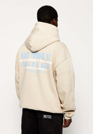Man draagt een beige hoodie met lichtblauwe en witte tekst "In the back of the bus" en zwarte broek met "MLTPLY Multiply Apparel" logo.