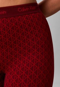 Røde leggings med et tekstureret mønster af overlappende diamanter og hjerter, med en kontrastfarvet rød talje, der viser "Calvin Klein"-mærket.