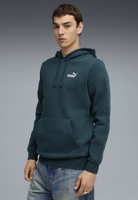 Sweat à capuche bleu canard en tissu doux, avec une poche kangourou, une capuche à cordon de serrage et un logo Puma blanc sur la poitrine, associé à un jean bleu délavé.