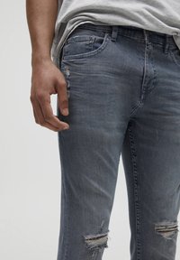 Jeans de denim azul desgastado con corte ajustado, que presentan detalles desgastados y rotos en las rodillas, botonera metálica y diseño de cinco bolsillos.