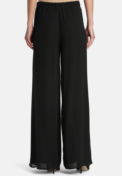 Personne portant un pantalon large noir ample avec une taille élastique, debout, les mains détendues le long du corps, vue de dos.