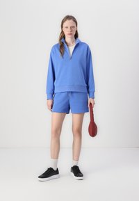Blauwe katoenen trui met halve rits, gecombineerd met bijpassende shorts. Kenmerkt zich door een geribde zoom en houdt een rood accessoire vast. Zwarte sneakers met witte zolen.
