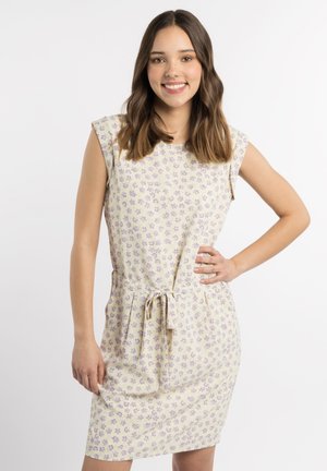 Junge Frau mit schulterlangem braunem Haar lächelt, sie trägt ein ärmelloses beiges Kleid mit kleinen lila Blumen und einem Taillenband.