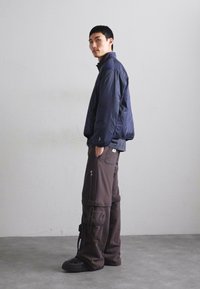 Giubbotto trapuntato blu navy con collo alto, abbinato a pantaloni cargo marroni larghi con cerniere e tasche. Calzature nere completano il look.