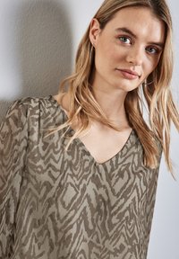 Durchsichtige, gemusterte Bluse in olivgrün mit V-Ausschnitt und langen Ärmeln, die ein dezentes Tierdruckdesign und eine weiche Textur aufweist.