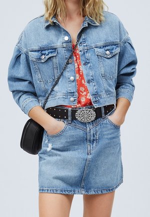 Woman wearing light blue cropped denim jacket and skirt, red floral blouse, black belt with ornate buckle, hands in pockets, and black crossbody bag.