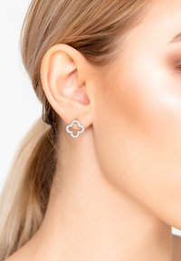 Orecchini a bottone in tono argento a forma di trifoglio, con piccoli cristalli trasparenti attorno ai bordi. Texture liscia, design minimalista.