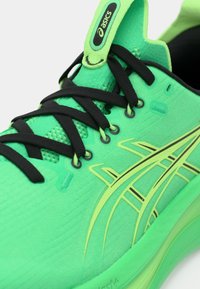 Sapatilha de corrida verde com um superior de malha texturizada, atacadores pretos e um design de destaque em verde claro. Possui uma sola almofadada e uma etiqueta de marca.