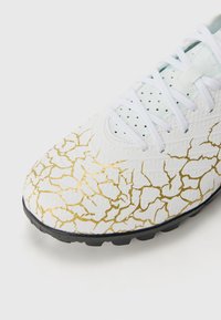 Chaussure de football blanche avec un motif doré fissuré, tige texturée, semelle noire et lacets blancs ; conçue pour l'adhérence et la performance.
