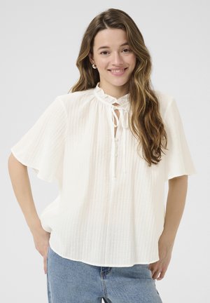White blouse with a ruffled collar, short flutter sleeves, and button closure. Textured fabric with a relaxed fit. Worn with blue jeans.