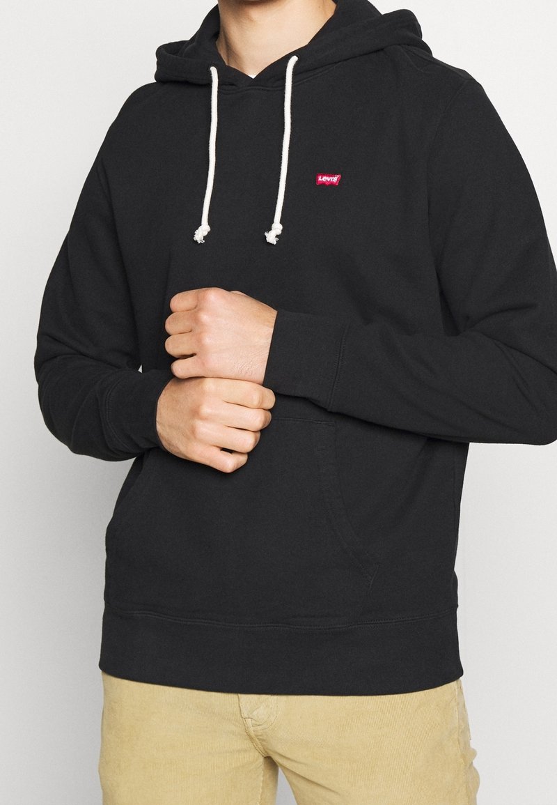 Personne portant un sweat à capuche noir Levi's avec des cordons blancs et un pantalon beige, les mains jointes au niveau de la taille sur un fond uni.