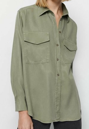 Personne portant une chemise boutonnée vert olive avec deux poches à rabat à l’avant et des manches longues, partiellement rentrée dans un pantalon sombre.