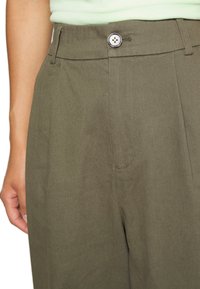 Pantaloni verde oliva con una texture liscia, dotati di chiusura frontale con bottone e tasche laterali; design sartoriale con una vestibilità rilassata.