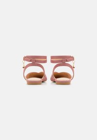 Sandalias de ante rosa con puntera cerrada, talón abierto, correas para el tobillo y herrajes en tono dorado. Suela plana con un detalle de textura mínima.