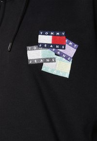 Sudadera negra de tela con capucha que presenta un parche bordado con rectángulos coloridos que muestran "TOMMY JEANS" y "TO JEANS" en varios colores.