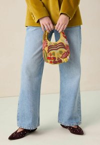 Sac à main en perles de couleur jaune avec un haut arrondi, présentant des motifs floraux en rouge et vert ; porté avec un jeans bleu clair et des ballerines foncées.