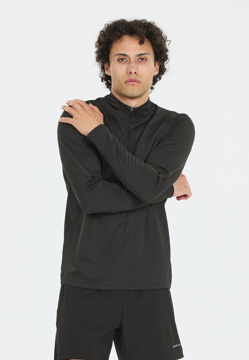 Schwarzes, langärmliges Sportoberteil mit hohem Kragen und Viertelreißverschluss; glatte Textur mit dezenten Nähdetails. Getragen mit schwarzen Shorts.