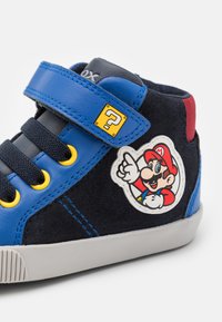 Blå och marinblå hög sneaker med en mocka-liknande textur, gula accenter, kardborreband och en Mario-karaktärspatch på sidan.