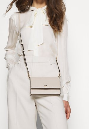 Vrouw die witte hoogtaillepantaloen en een witte blouse met een strik draagt, met een beige Michael Kors schoudertas.