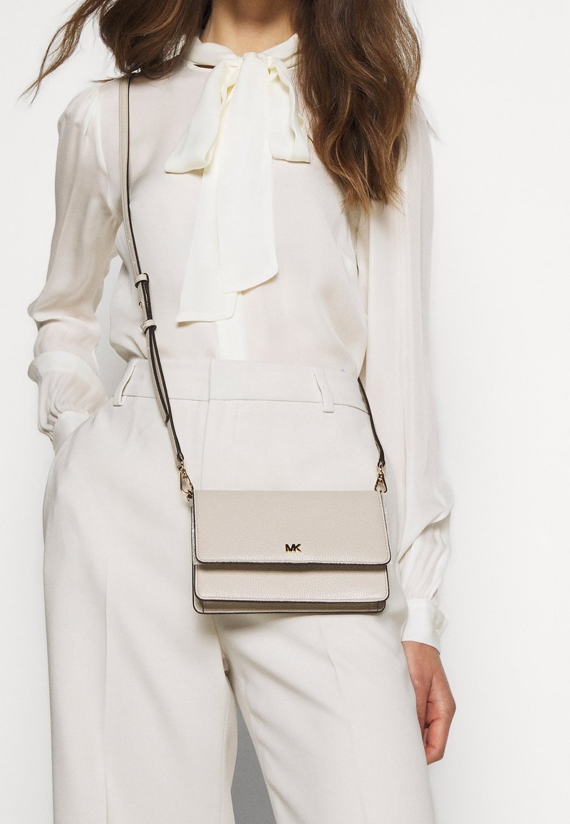 Femme portant un pantalon blanc taille haute et un chemisier blanc avec un nœud, portant un sac bandoulière beige Michael Kors.