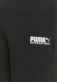 Sorte joggingbukser lavet af blødt stof med en hvid "PUMA INTERNATIONAL"-logo på venstre lår.