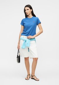 Γυναίκα που φορά μπλε μπλουζάκι, λευκά σορτς μέχρι το γόνατο, ανοιχτό μπλε πουλόβερ δεμένο στη μέση, μαύρα σανδάλια, κρατώντας μικρό μαύρο τσαντάκι.