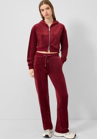 Bordeaux farbener Velours-Trainingsanzug mit einer kurzes Zip-Hoodie und passenden Hosen, ausgestattet mit einem Kordelzugbund und einer weichen, glatten Textur.
