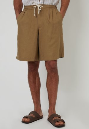 Pantalones cortos de lino marrón con cintura elástica y cordón. Con bolsillos laterales y un corte relajado, combinados con sandalias de dedo marrones.