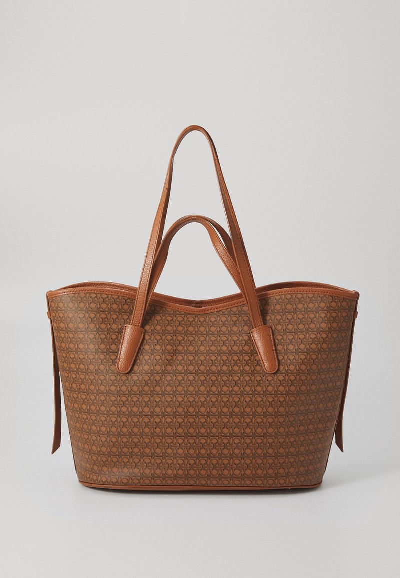 Sac cabas en cuir marron avec surface à motifs, deux longues bandoulières et deux poignées plus courtes sur fond clair uni.