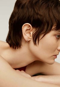 Pendiente de aro de plata con un pequeño acento de bola. Primer plano de un perfil lateral resaltando la textura del cabello y el tono de piel. Fondo neutral.