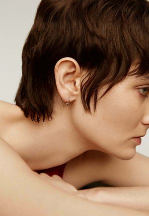 Pendiente de aro de plata con un pequeño acento de bola. Primer plano de un perfil lateral resaltando la textura del cabello y el tono de piel. Fondo neutral.