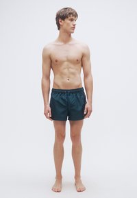 Μαγιό βερμούδα σε σκούρο πράσινο χρώμα, κατασκευασμένο από λείο ύφασμα με ελαστική μέση και κορδόνι ρύθμισης. Διαθέτει πλαϊνές τσέπες και λεπτομέρεια λογότυπου.