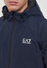 Mørkeblå vandtæt jakke med snor i hætten, lynlåslukning og hvid EA7-logo på venstre bryst. Glat tekstur med minimalistisk design.