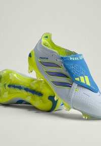 Chaussures de soccer Adidas Predator blanches et jaune néon, une chaussure inclinée montrant les crampons de la semelle, avec un couvre-lacets bleu affichant le logo Adidas et le texte "EST. 1994".