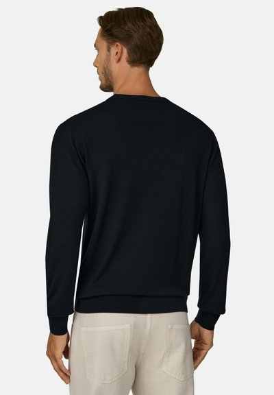 Homme aux cheveux courts bruns, vu de dos, portant un pull noir à manches longues et un pantalon beige clair avec des poches arrière.
