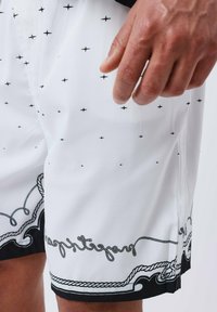 Bañadores blancos con pequeños patrones de cruces negras, con un borde decorativo en negro y texto en un diseño de cuerda en el dobladillo.