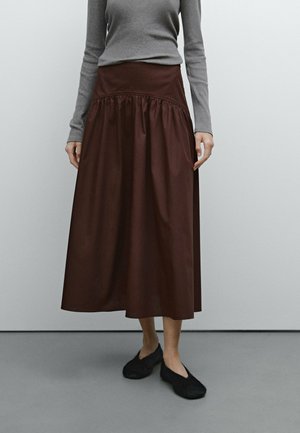 Woman wearing a brown midi skirt, gray long-sleeve fitted top, and black flat shoes, standing against a plain light gray background.