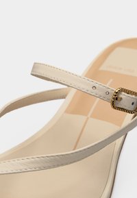 Κοντινό πλάνο μπεζ σανδάλι με λεπτές δερμάτινες λουριά και μικρή χρυσή αγκράφα σε ουδέτερο φόντο.