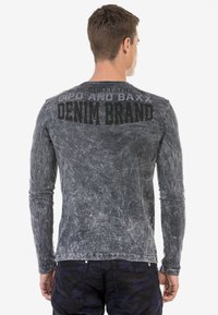 Langärmliges graues T-Shirt mit einem verwaschenen, strukturierten Finish. Auf der Rückseite befindet sich schwarzer Drucktext, der die Phrasen "GIPO AND BAXX" und "DENIM BRAND" umfasst.