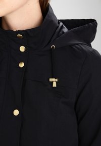 Svart huva jacka gjord av slät tyg, med gyllene knappar, en dragsko med justering, och två bröstfickor med platta sömmar.