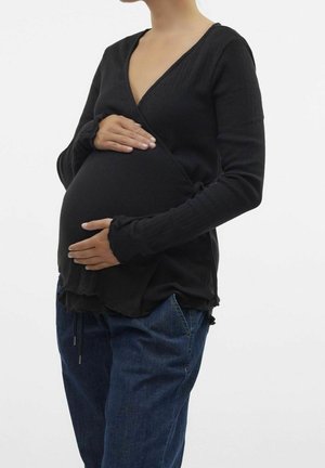 Osoba w ciąży ubrana w czarną bluzkę z długim rękawem i niebieskie dżinsy, delikatnie trzymająca brzuch obiema rękami na jednolitym tle.