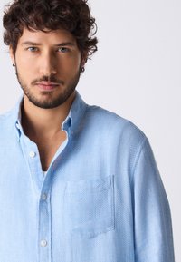 Hele-sinine, pika varrukaga särk tekstuurse kangaga, millel on nööbitav krae ja rinnatasku. Materjalis on peen muster.