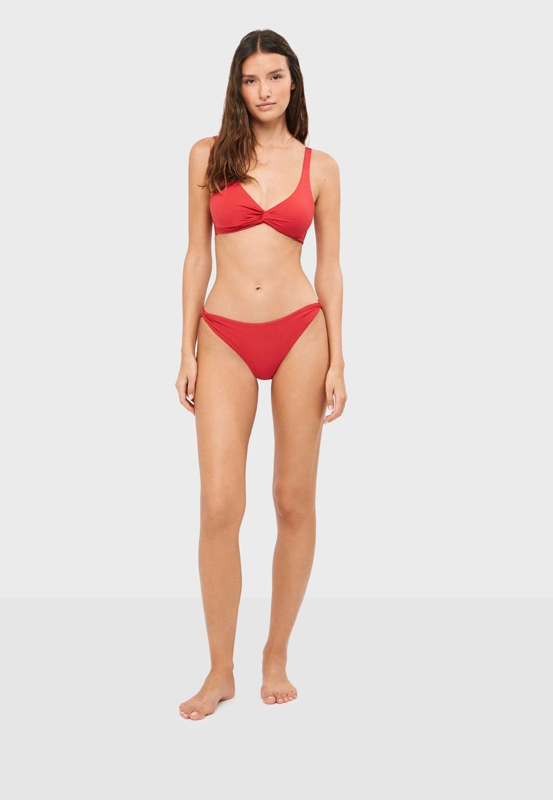 Femme debout pieds nus portant un maillot de bain deux pièces rouge avec un haut torsadé devant sur un fond clair uni.