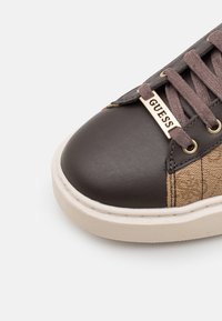 Baskets marron et noires avec un bout en cuir lisse, des panneaux latéraux texturés couleur sable, des détails dorés du logo et des lacets marron.