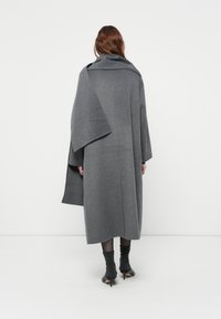 Vrouw die met de rug naar de camera staat, gekleed in een lange grijze jas met een ruime scarf en zwarte doorschijnende panty’s en hakken.