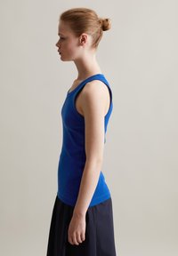 Niebieska, dopasowana bluzka na ramiączkach wykonana z gładkiego materiału, z szerokimi ramiączkami, zestawiona z ciemną spódnicą, prezentowana w profilu bocznym.