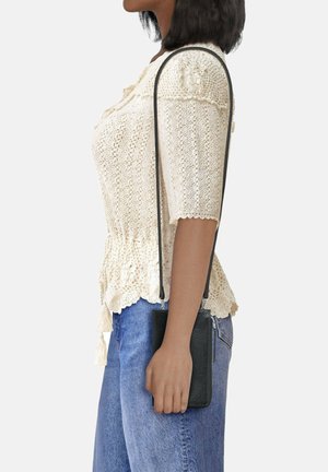 Schwarze, strukturierte Leder-Crossbody-Tasche mit langem Trageriemen, gehalten in einer Hand, kombiniert mit einem leichten Strickoberteil und blauen Jeans, seitlich gezeigt.