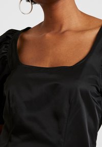 Czarna satynowa bluzka z kwadratowym dekoltem, bufiastymi rękawami i gładką fakturą. Widoczne detale szwów wzdłuż szwów.